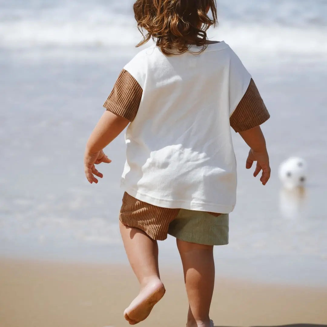 Child walking on a beach wearing a white shirt with brown sleeves and shorts. Organic cotton corduroy shortd for kids toddlers and infants