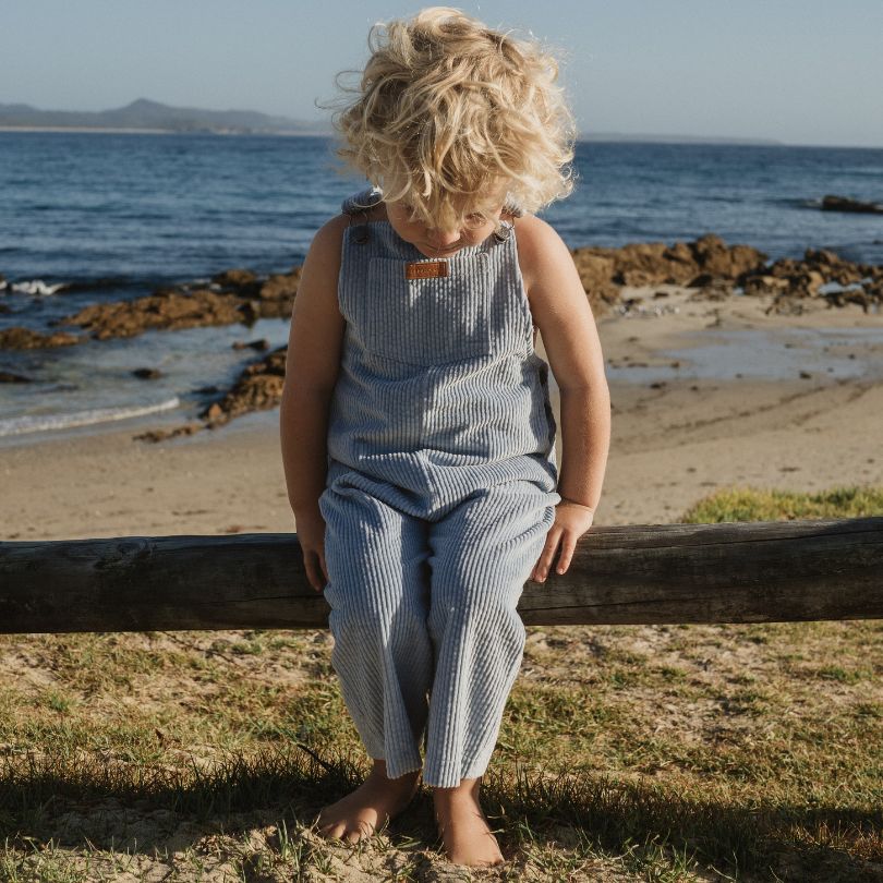 boy sitting on a fence at the beach wearing his Kids and toddlers blue corduroy overalls or dungarees