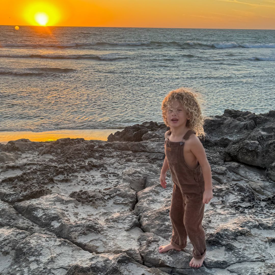 girl wearing her brown Kids and toddlers corudoy overalls or dungarees on some rocks by the beach