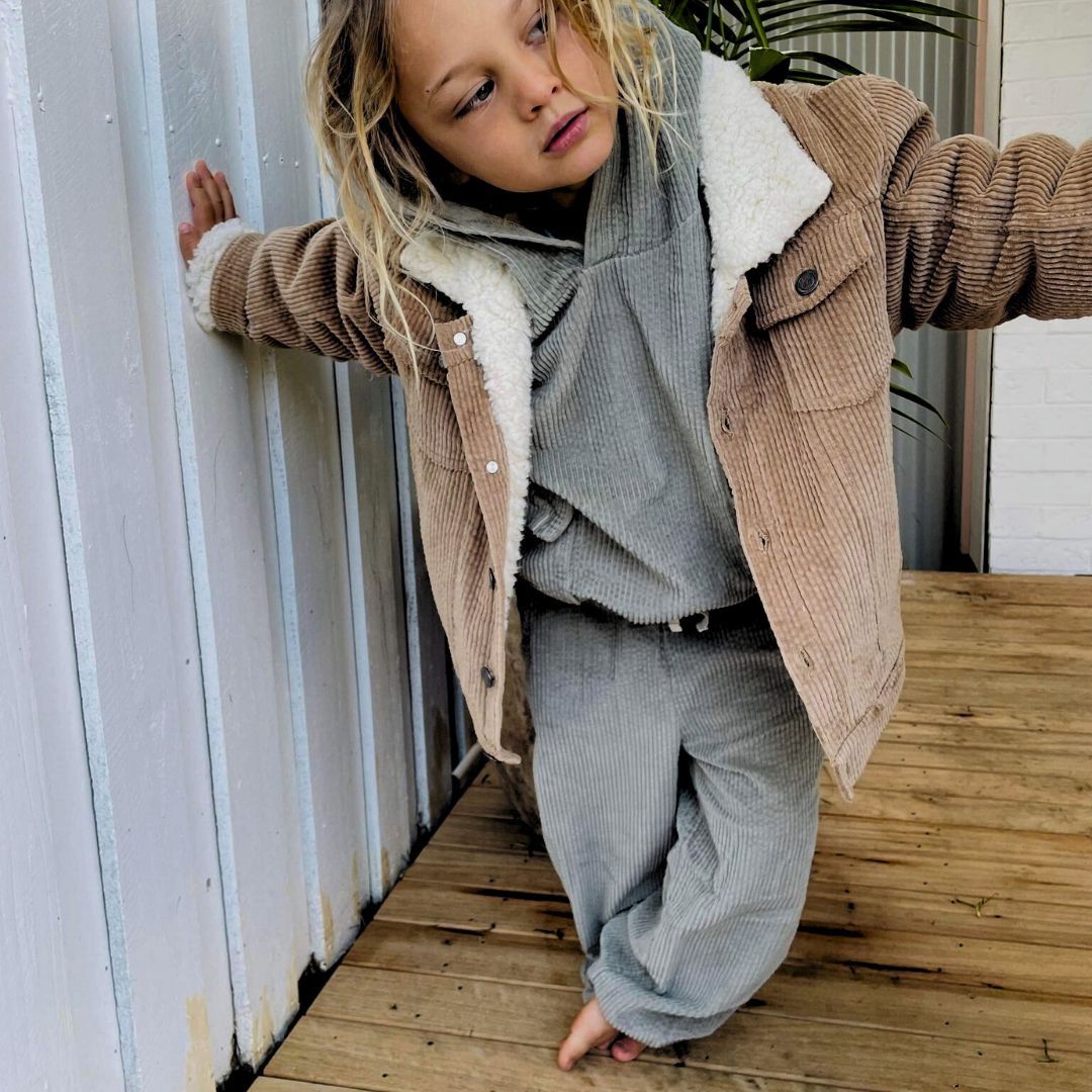 a boy wearing a Kids and toddlers organic corduroy sherpa fleece jacket in tan with a sage corduroy tracksuit underneath standing on a deck with one hand on a fence