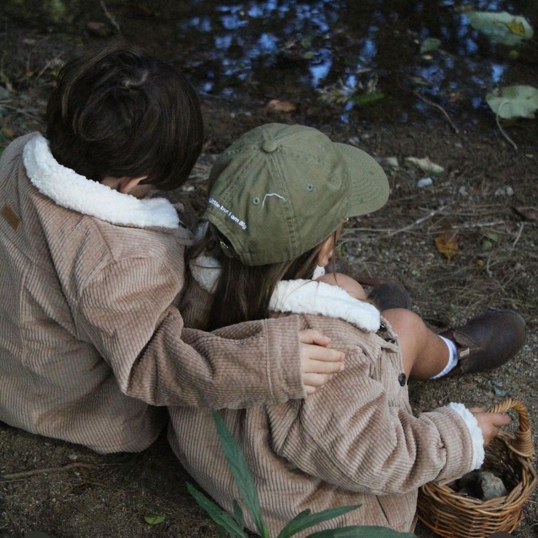 Kids and toddlers wearing a kids organic cotton corduroy sherpa fleece jackets sitting down on the ground near a river